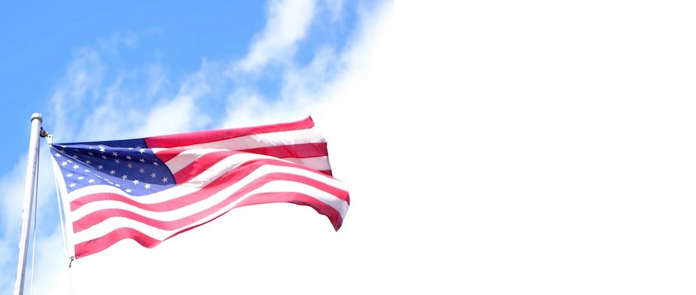 An American flag waves in the wind against a blue sky with some wispy clouds, symbolizing freedom and unity. The flag's red and white stripes and white stars on a blue field are clearly visible. The right third of the image is mostly blank with a white background, evoking thoughts of ICE Enforcement Priorities.