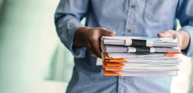 A person in a blue shirt is holding a large stack of files and documents, organized meticulously with black and orange binder clips. The background is blurred, drawing focus to the files and the person's hands, possibly a member of Lancaster Immigration Lawyers assisting with case preparations.