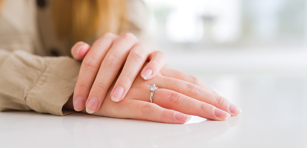 A close-up of a person's hands resting on a white surface. The person is wearing a light-colored shirt, and there is a diamond engagement ring on their left ring finger, possibly seeking assistance from Lancaster Immigration Lawyers. The background is softly blurred.