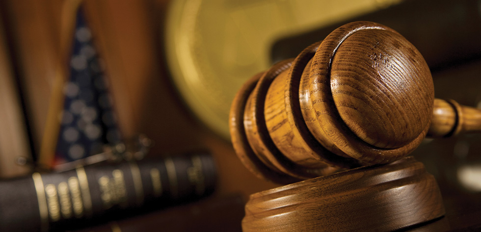 Close-up of a wooden gavel resting on a sound block in a courtroom scene. In the blurred background, a portion of the United States flag and some legal books are visible, suggesting a setting in a court of law where Lancaster Immigration Lawyers might advocate for justice and legal proceedings.