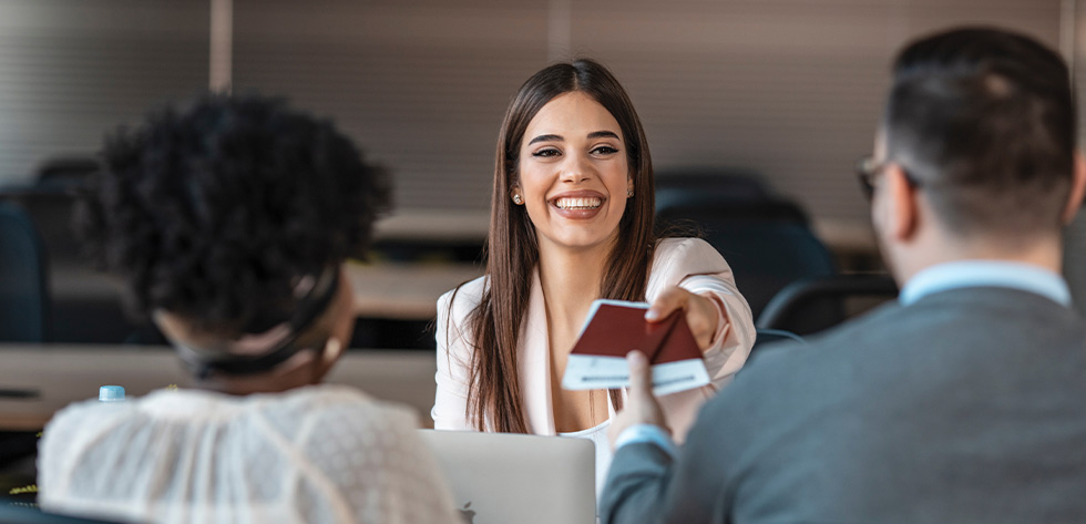 A woman with long brown hair is smiling and handing a passport to a man wearing glasses. Another woman with curly hair and a headband is seated nearby. In the modern office of Lancaster Immigration Lawyers, laptops and papers are scattered on the table.