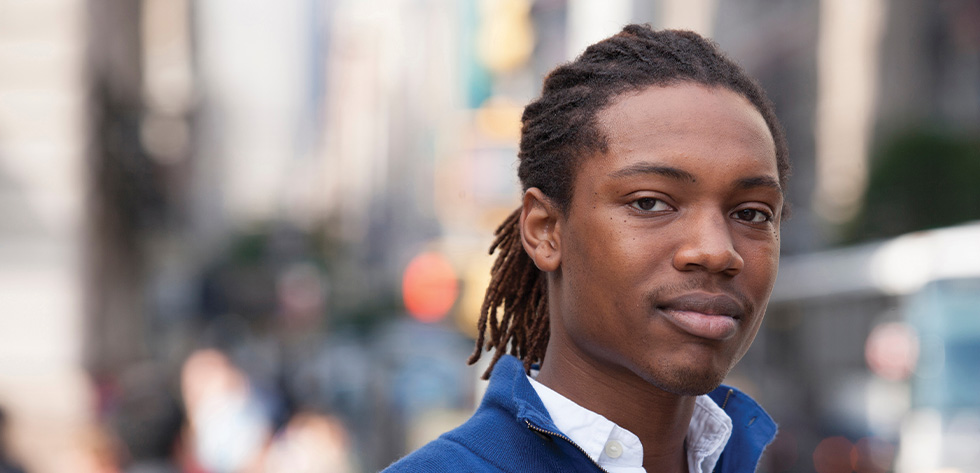 A person with long hair in locs is standing outdoors in an urban setting, wearing a blue jacket over a white shirt. The background is blurred, showing busy city life with buildings and people, perhaps on their way to consult Lancaster Immigration Lawyers.