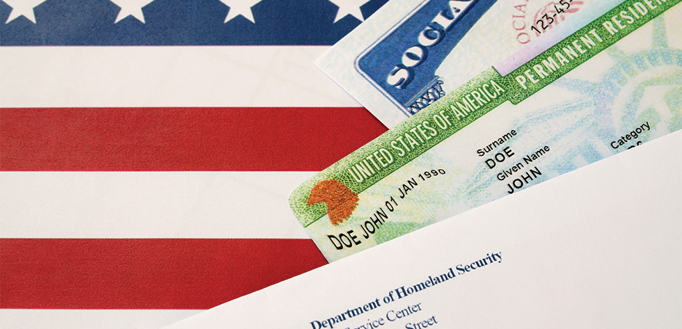 A close-up of a U.S. flag partially covering documents including a Social Security card, a Permanent Resident Card, and a letter from the Department of Homeland Security. The Permanent Resident Card shows the name 