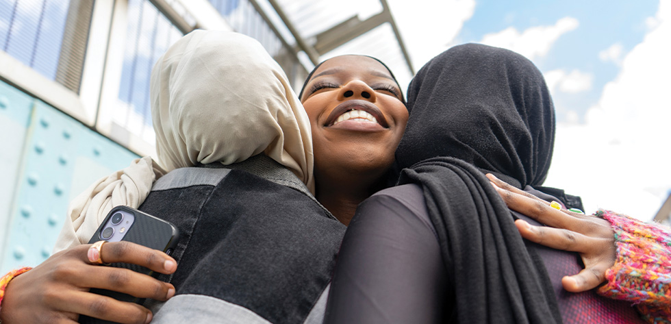 Three people share a warm embrace. The person in the middle, smiling brightly, is surrounded by the other two whose backs are turned to the camera. One of them holds a smartphone. In the background, a building and a partly cloudy sky frame their happiness, possibly celebrating good news from Lancaster Immigration Lawyers.